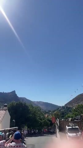 The video shows a bright sunny day with a clear blue sky. The sun is prominently visible in the upper left corner, casting a strong glare. In the background, there are mountains covered in trees. In the foreground, a line of people is visible, walking down a street lined with buildings. Some of the buildings have tile roofs. Small colorful flags are strung across the street in the distance. The camera appears to be held at eye level, and there is a brief moment where a finger enters the frame in the lower right corner.
