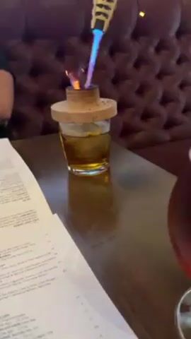 The video shows a hand holding a torch lighter with a blue flame burning the wood of a cocktail smoker placed on top of a glass of amber-colored liquid. A menu and another glass of amber liquid are also on the dark brown wooden table. The background is a tufted burgundy leather booth seat.