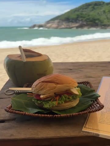 A table made of rough wood holds a burger, a coconut with a straw, and a partially seen menu. The burger sits on a woven tray lined with a large leaf. Beyond the table is a sandy beach leading to a turquoise ocean with waves, and a green, hilly headland extends into the sea. The view from beneath an umbrella is toward a partly cloudy blue sky.