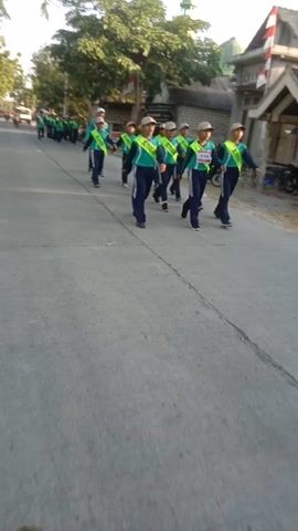 The video shows a group of people in uniform marching on a street. The uniforms consist of a light blue shirt, a green vest, and dark pants. They are all wearing hats. The street is paved and appears to be in a rural or suburban area, with trees and buildings visible in the background. The camera is moving, giving a sense of motion. One person in a green shirt and dark pants is walking in front of the group. Another person on a motorcycle is also visible. The lighting suggests it is daytime, possibly late afternoon.