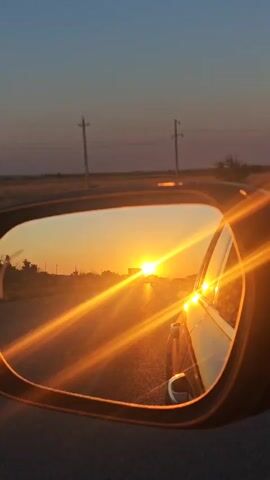 A car's side mirror reflects a vibrant sunset with the sun low on the horizon casting a golden glow. The reflection shows a road, power lines, and sparse vegetation. The car's side is visible in the reflection, with water droplets on the mirror. The sky transitions from a deep blue to orange and yellow hues as the sun sets.
