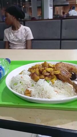The video shows a close-up view of a meal on a green tray. The meal consists of rice topped with fried plantains and chicken. A bottle of water is also on the tray. In the background, a young woman wearing a headscarf and a light-colored shirt is seated at a table. The environment appears to be a food court or restaurant, with other tables and people visible in the distance. The camera pans slightly, revealing more of the table and the person seated in the foreground, who is wearing a purple garment. The lighting is dim, suggesting an indoor setting at night or in the evening.