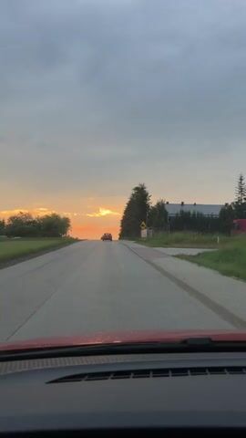 The video shows a view from inside a car driving on a road towards a sunset. The sky is overcast with streaks of orange and yellow from the setting sun. There are trees on the right side of the road and some buildings in the distance. Two cars are visible on the road ahead, one slightly further than the other. The car in front is a dark color and the car further down the road is lighter in color. The camera is looking forward through the windshield, and the dashboard of the car is visible at the bottom of the frame. The overall impression is of a peaceful evening drive.