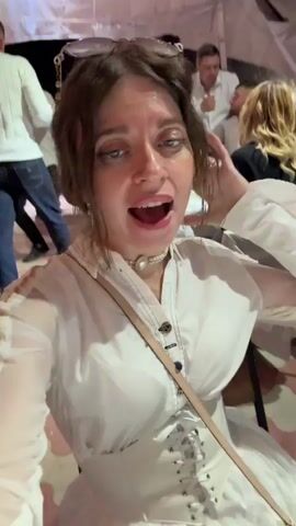 A woman with brown hair and bright makeup is filming herself in the foreground. She is wearing a white shirt with intricate details and a pearl necklace. Behind her, a group of people are dancing and socializing under a tent with string lights. The floor is a patterned pink and white tile. The atmosphere appears festive and lively, possibly at a wedding or party.