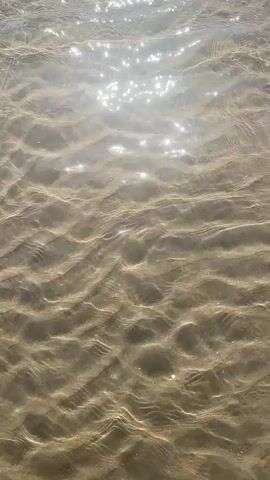 The video shows a top-down view of shallow, clear water flowing over a sandy bottom. The sand has distinct ripples and patterns. Sunlight reflects off the surface of the water, creating shimmering highlights. The water appears calm, with gentle movement disturbing the sand. No people or other objects are visible.