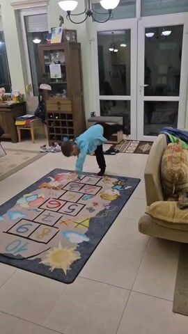 A young boy is indoors in a brightly lit room that appears to be a living or family room, doing acrobatic movements on a hopscotch rug. He performs a handstand, and then transitions to a full split on the rug. He smiles at the camera while in the split. The room contains furniture including a sofa, display cabinet, and a small table. There are shoes scattered on the floor, and a doorway leads to an outdoor area or another room.
