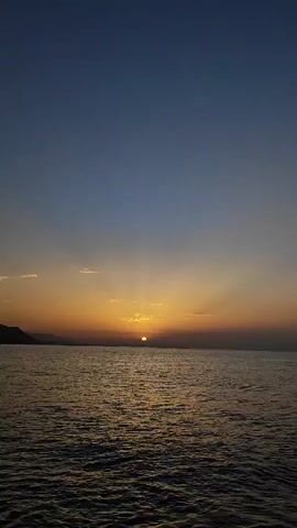 A video captures a sunset over the ocean. The sun is partially visible, casting a golden glow across the horizon. The sky transitions from a deep blue at the top to shades of orange and yellow near the horizon. Rays of light emanate from the sun, creating a dramatic effect. The ocean surface is rippled, reflecting the colors of the sunset. In the distance, a dark silhouette of land is visible. The overall scene is serene and depicts the beauty of nature at dusk.