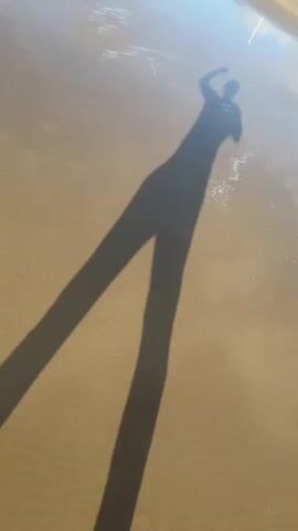 A person's shadow is cast on wet sand. The shadow is long and distorted, with one leg extended forward. The shadow appears to be moving. The sun is shining, creating the shadow.