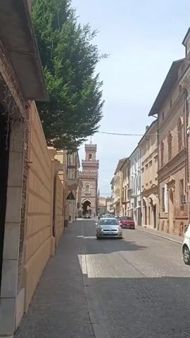 The video shows a view down a cobblestone street in a town. On the left, there is a beige wall with horizontal brown stripes, a building with some decorative string lights, and a large tree. On the right, there are several buildings with arched windows and red tile roofs, typical of Italian architecture. In the distance, a tall tower with a clock and an arched base is visible. Several cars are driving down the street, including a silver hatchback in the foreground. The sky is bright and overcast.