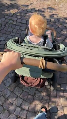 The video shows a first-person perspective of someone pushing a stroller on a cobblestone path. The stroller contains a young child with blonde hair, facing away from the camera. The child is wearing a light-colored t-shirt and is secured in the stroller with a harness. The stroller itself is green with brown leather accents on the handle. A brown bag is visible hanging from the back of the stroller. Sunlight casts shadows on the path, suggesting it is daytime. The scene appears to be outdoors, possibly a park or a residential street, given the cobblestone and dappled sunlight. The overall mood seems peaceful and leisurely, likely a walk in the park. There is no visible text in the media. The location context of Bad Sobernheim, Germany, suggests a European setting.