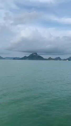 The video shows a view of the sea from a boat. The sea is a turquoise color and there are islands in the distance. The sky is cloudy and gray. The video appears to be shot from a moving boat, with the horizon gradually changing. There is speech heard in the background.
