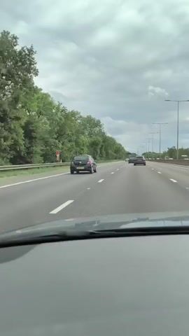 The video shows a view from inside a car driving on a highway. The sky is cloudy. There are other cars on the highway, some in front and some to the side. Trees and bushes line the side of the highway. There are streetlights along the road. The dashboard of the car is visible in the foreground.