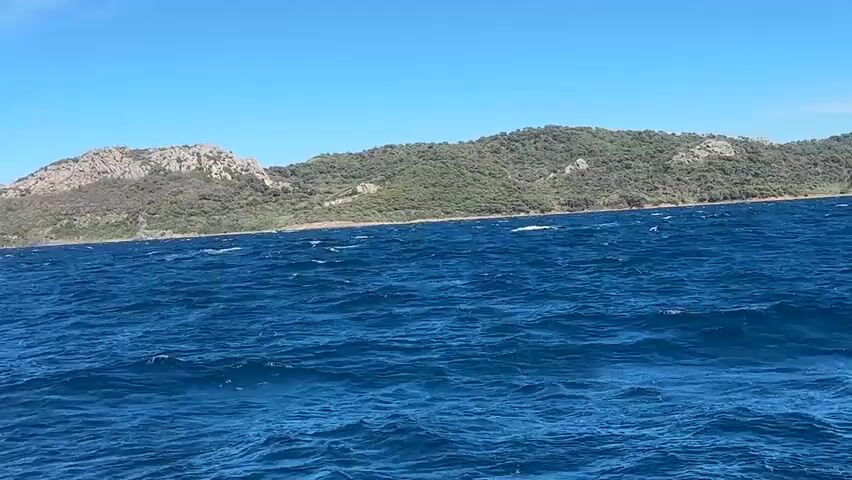 The video shows a choppy blue ocean under a clear sky. In the distance, a green, hilly island with rocky outcrops is visible. The camera appears to be mounted on a moving boat, as evidenced by the white railing in the lower left corner and the spray of water. The water is rough, with waves and whitecaps. The sky is bright blue with no clouds. The overall scene suggests a sunny day at sea.
