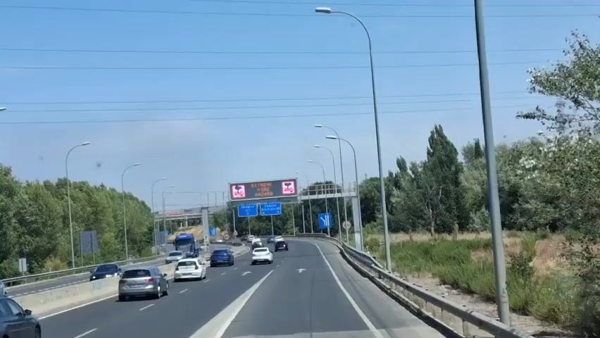 The video shows a daytime view from a vehicle traveling on a highway. The sky is clear blue with some power lines visible. The highway has multiple lanes and is carrying a moderate amount of traffic, including cars and trucks. On the right side of the highway, there is a grassy embankment with trees and vegetation. On the left side, there are more trees and some buildings or infrastructure in the distance. Above the highway, there is an electronic road sign displaying "EXTREME FIRE HAZARD" in English, with accompanying icons of flames and a fire extinguisher. There are also blue directional signs for upcoming exits or route information. The overall impression is a sunny day on a busy highway with a warning about fire risk.