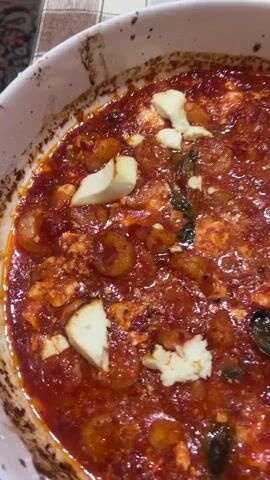 A close-up overhead shot shows a white ceramic baking dish filled with a bubbling red sauce. Scattered throughout the sauce are small, pink shrimp and chunks of white feta cheese. Dark green basil leaves are also visible, and the edges of the dish appear slightly browned, suggesting it has been baked. The dish rests on a table covered with a checkered tablecloth. To the right of the dish, a jar filled with green pickles is partially visible.