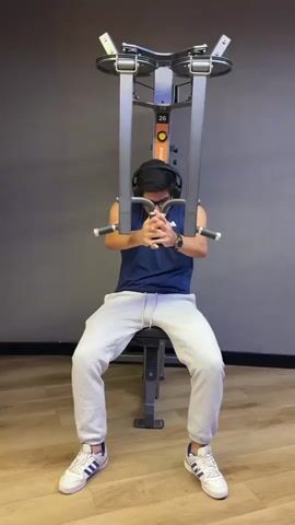 A young man is performing pec fly exercises on a gym machine in an indoor gym setting. He is seated, wearing a navy blue sleeveless tank top, light grey sweatpants, and white sneakers with blue stripes. He also wears glasses and black over-ear headphones.

The man executes the exercise by extending his arms wide to grip the machine's handles, then bringing them together in front of his chest, engaging his pectoral muscles. His facial expressions indicate concentration and effort during the movement.

The background consists of a dark grey wall and a light wood-patterned floor. The gym machine, possibly a pec deck, is silver and grey with an orange accent on its central column. The number "26" is clearly visible on the orange section of the machine, along with the brand name "OROCO" below it. This scene is set in a gym in San Isidro, Peru.