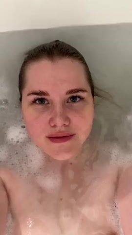A woman is shown from an overhead angle in a bathtub full of water and white bubbles. The woman opens her eyes, looks directly at the camera, closes her eyes, and smiles. The bathroom is white.