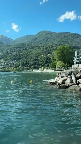 The video shows a bright, sunny day at a lake. The foreground is dominated by rippling blue-green water. In the middle ground, several yellow buoys are visible in the water. People are seen in kayaks or on paddleboards in the lake. The background features a lush green, tree-covered mountainside with some buildings visible. To the right of the image, a rocky shore leads up to a white railing and a glimpse of a structure. The sky is a clear, vibrant blue with a few wispy white clouds.