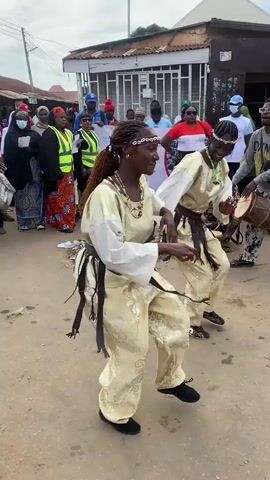 Two women in traditional attire are dancing in the middle of a dirt road, with a crowd of people watching them. The dancers are wearing long, flowing robes and beaded jewelry. One of the dancers is playing a drum. In the background, there is a banner with text on it, and several buildings. The overall scene appears to be a cultural celebration or event.