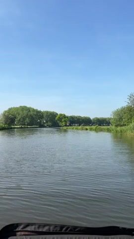The video shows a view from a moving boat on a waterway. The water is gray and rippled, reflecting the sky. On both sides of the waterway are trees and shrubs, forming a green border. The sky is blue with some scattered clouds. There is no visible person in the video. At one point, what appears to be a traditional windmill is visible through the trees. The video appears to be shot during daylight hours in a natural setting.