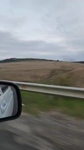 The video captures a view from inside a moving vehicle, looking out past the passenger side mirror towards a rolling landscape. The mirror reflects the interior of the car, showing a portion of the dashboard and a white car door with a red accent light. The foreground outside the car shows a highway guardrail with green vegetation visible beneath it. Beyond the guardrail, vast, dry, brown fields stretch across gently sloping hills. The sky is overcast with a soft, diffused light, suggesting it might be early morning or late afternoon. The overall scene conveys a sense of travel through a rural, open area.