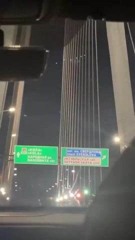 The video is a first-person perspective from inside a vehicle at night. The vehicle is moving over a bridge, as evidenced by the visible support cables and roadway lighting. Two green road signs are visible through the windshield. The sign on the left indicates "M-11 KOLÀ" and "NARODNAYA str." with an arrow pointing left. The sign on the right indicates "pos. im. SVERDLOVA," "OKTYABRSKAYA emb.," and also has an arrow pointing left. The background shows a dark sky and the illuminated vertical lines of the bridge's suspension system. There are also streetlights and vehicle headlights visible on the road ahead. The interior of the car is dimly lit, with a rear-view mirror visible at the top of the frame. The overall impression is of traveling through a city at night on a large bridge.