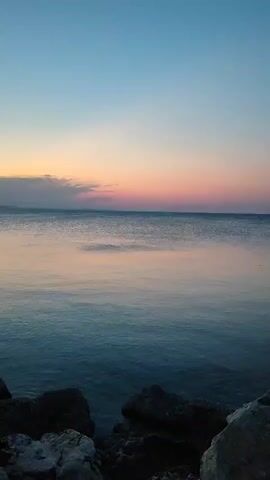 A video displays a calm ocean under a clear sky at sunset. The sky transitions from a soft blue at the top to hues of pink, orange, and yellow near the horizon. The water reflects the colors of the sky, creating a serene and peaceful atmosphere. Rocks are visible in the foreground along the shoreline.