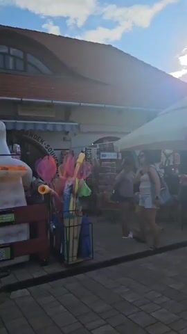 The video shows a street scene on a sunny day. There are people walking around, some are sitting at tables under umbrellas, and there are bicycles parked. The buildings in the background appear to be shops and restaurants. The overall atmosphere is relaxed and casual, suggesting a tourist area or a local gathering place. The sun is shining brightly, casting long shadows on the paved ground. The sky is blue with some scattered clouds. There's a variety of merchandise displayed outside a shop, including colorful items that look like beach toys or decorations. Several people are walking away from the camera, some carrying bags. A church steeple is visible in the distance.