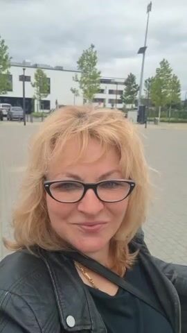 A selfie video shows a woman with blonde hair and glasses smiling at the camera. She is wearing a black leather jacket over a dark shirt and a gold necklace. In the background, there are modern buildings under construction, trees, and streetlights. The sky is overcast.