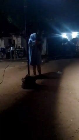 The video shows an outdoor scene at night, likely in a village or rural area. The area is dimly lit, with a person standing near a microphone stand in the foreground. A few people are sitting at tables in the background. Some people are standing behind the sitting people. A white tent is visible as well. A sound system is heard, suggesting there may be a performance or public gathering taking place. The ground appears to be dirt or sand.