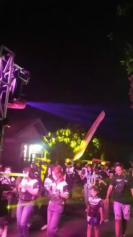 The video captures a lively outdoor event at night in Dukuh Klopo, Indonesia. A group of people, including adults and children, are gathered, seemingly enjoying a celebration or party. The scene is illuminated by a dynamic light show featuring vibrant purple, green, blue, and red beams, likely from stage lighting and possibly laser effects.

Several individuals are visible in the foreground and background, some appear to be dancing or socializing. Some people are wearing matching t-shirts with green and white accents, suggesting they might be part of a group or team. One child in the foreground is wearing a dark t-shirt with a graphic design.

The setting appears to be an open area, possibly a street or a yard, with trees and a building with illuminated windows visible in the background. The overall atmosphere is festive and energetic, indicated by the use of colorful lights and the presence of a crowd. The time of day is clearly night, and the weather appears to be clear.

There is some text visible on what appears to be a speaker or stage setup in the background. In frames 00:18 and 00:19, the text "SEKTOR BARAT" is discernible. The event seems to be a public gathering, possibly a local festival or a community event, characterized by music and vibrant light displays.