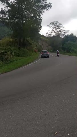 A black car turns a corner on a winding road. A motorcycle is visible in the distance. Greenery and hills surround the road. The sky is overcast.