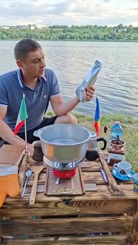 A man crouches beside a wooden table set up outdoors by a body of water. He is holding a package in his right hand and appears to be speaking to the camera. On the table, there is a pot of water on a portable stove, a coffee grinder, a moka pot, and two small flags, one of Italy and one of Romania. The background shows a calm body of water with trees on the far bank and a cloudy sky.