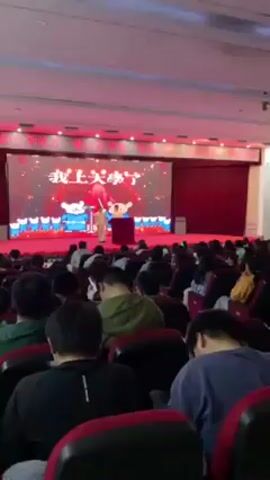 The video shows a large indoor audience seated in rows of red chairs, facing a stage. On the stage, a person dressed in traditional Chinese clothing stands behind a podium. A large screen behind the person displays Chinese characters and animated cartoon figures. A firework display erupts behind the person on stage. The audience is diverse, with a mix of people of different ages and clothing styles. Many individuals are using their phones, possibly recording the event. The room is well-lit with a combination of overhead and stage lighting.