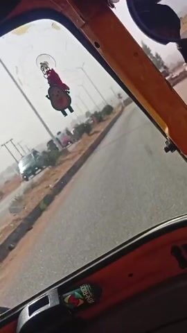 A low-angle shot from inside a moving auto rickshaw shows a straight road lined with street lights. There is a pink keychain attached to the windshield. A man in a plaid shirt is walking near a barrier of sandbags on the right side of the road. Several cars and another auto rickshaw are visible in the distance. The sky is overcast.