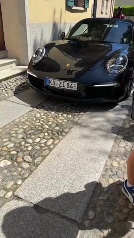 The video shows a black convertible Porsche parked on a cobblestone street. The car has a German license plate that reads "KA ZI 84". A motorcycle passes in the background. Two women, one wearing a red and white checkered hat, stand nearby. The camera then pans to reveal a view of a lake surrounded by trees. There are tables and chairs set up near the water's edge. The scene appears to be in a European town on a sunny day.