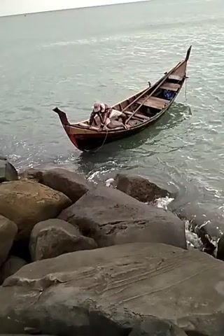 The video shows a small wooden boat in the water near a rocky shore. A person is standing in the water next to the boat, appearing to be working on it or preparing to leave. The sky is overcast and the water is relatively calm. The boat is of a traditional design, with a raised bow and stern. The rocks on the shore are large and dark. The overall scene suggests a coastal or marine environment with fishing activities.