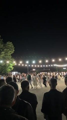 A video captures a nighttime outdoor gathering where a group of people are holding hands and dancing in a circle. The scene is illuminated by strings of bare bulbs hanging overhead, casting a warm glow on the participants and the surrounding dark environment. Trees are visible on the left side of the frame, and in the background, structures that appear to be buildings or stalls are faintly lit. The foreground shows the backs of several individuals watching the dance, with their faces obscured. The overall atmosphere suggests a festive community event or celebration.