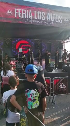 Certainly! Here's a detailed description of the scene in the video:

The video shows a live musical performance at an outdoor event titled "Feria Los Altos," as indicated by the banner above the stage. 

A band consisting of several musicians is performing on a stage equipped with lighting and a large LED screen displaying a logo. Musicians are playing instruments like saxophone, trombone, drums, and guitar. 

Several people, including women and children, are standing in front of the stage, watching the performance. One child wears a blue baseball cap and a black shirt with a panda graphic on the back. 

The area is divided by red and silver metal barriers. The overall atmosphere appears to be that of a fair or festival with live entertainment.