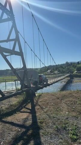 A dark colored SUV and a trailer are crossing a narrow suspension bridge over a river. The bridge appears to be constructed of wood and metal. The SUV is in the lead, followed by the trailer. The sun is shining brightly, and the sky is clear blue. The bridge is surrounded by trees and hills. The river is flowing rapidly below the bridge. The camera is positioned on the bank of the river, looking up at the bridge. The video is shaky, suggesting it was filmed handheld.