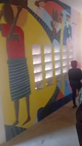The video shows a long mural on a wall inside what appears to be a school building. The mural is brightly colored with depictions of people carrying items on their heads and silhouettes of figures. The mural is painted around a set of small, rectangular windows. People are seen walking down the hallway in the background. The wall seems to be adjacent to a corridor or hallway.