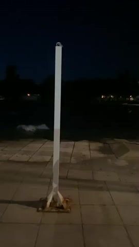 The video shows a dimly lit outdoor scene at dusk or night. A white pole stands in the foreground, casting a shadow on the paved ground. Beyond the pole, a body of water is visible, with distant lights reflecting on its surface. Silhouetted trees and land formations are discernible in the background. The camera pans slowly to the left, revealing a line of similar white poles, one of which is illuminated by a streetlamp, casting a warm glow on the walkway. The scene suggests a waterfront promenade or pier at the end of the day.