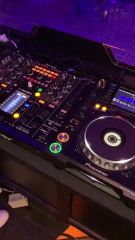 The video shows a close-up, high-angle shot of a DJ setup in a dimly lit room with purple and orange lighting. The equipment includes a Pioneer DJM-2000 mixer and a Pioneer CDJ-2000nexus CD player. The mixer has numerous illuminated buttons, faders, and knobs. The CD player has a large jog wheel with illuminated rings. A hand is briefly visible operating some controls on the mixer. The background appears to be a seating area with tables and chairs, possibly a lounge or club. The overall impression is of a professional DJ setup ready for use.