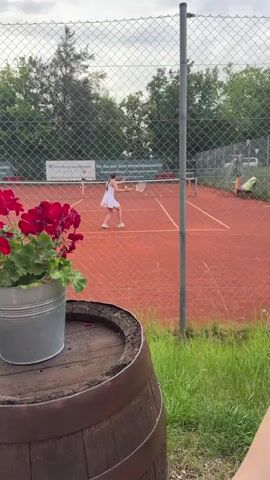 A young girl in a white tennis outfit is playing tennis on a red clay court. She is positioned on the left side of the court, facing the net and the other side where another player is visible in the distance. She is in the process of hitting a tennis ball with her racket. The court is enclosed by a chain-link fence. In the foreground, a wooden barrel with a metal pot of red flowers is visible. The background shows trees and a cloudy sky.