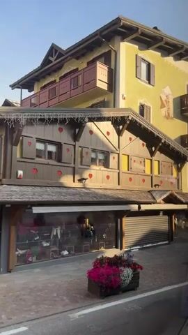 The video pans from left to right along a street in what appears to be a European town. On the left is a multi-story building with a light green facade. It has several balconies with dark brown railings. A mural of a figure in traditional clothing is visible on the upper part of the facade. Below the balconies, there are wooden shutters decorated with red heart shapes. The ground floor of this building appears to be a shop with large display windows, some of which are covered by rolling metal security gates. In front of the building is a wooden planter box filled with colorful flowers, predominantly pink and purple.

As the camera moves right, the building continues, with more balconies adorned with cascades of red, pink, and white flowers. There are signs visible, including one that says "GHEZZI SPORT" and another with a red oval logo that reads "Andel SPECK". Further along, there is an outdoor seating area for a restaurant or cafe with white canopies and tables. Another sign reads "CAMPBOANS".

To the right of this building, the street continues with more buildings, some of which are made of wood and have multiple levels of balconies also decorated with flowers. A pedestrian street is visible, with a man standing and looking at his phone. There are also flower planters along this street. The pavement is paved with stone. The sky is clear and blue, indicating daylight. The video appears to be shot from a moving vehicle, possibly a car, as indicated by the brief appearance of a car at the end of the clip. The overall impression is of a charming, well-decorated alpine town.
