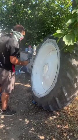 A person is spray painting a white wheel on a large tractor tire outdoors. The person is wearing a black t-shirt, grey shorts, and a grey bandana around their neck, with a mask covering their nose and mouth. They are holding a spray paint can and a spray gun. The tractor tire is lying on its side on the ground, with the white wheel prominently visible. The background shows trees and foliage, suggesting a rural or outdoor setting. There are also some cans and other items on a surface behind the person. The lighting indicates it is daytime.