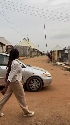 The video shows a woman with long braids walking along a dirt road in a rural area. She is wearing a white shirt and khaki pants. A silver car is parked on the side of the road. There are small houses and buildings in the background. A man walks down the street carrying a long pole and a motorcycle passes her in the opposite direction. The sky is overcast.