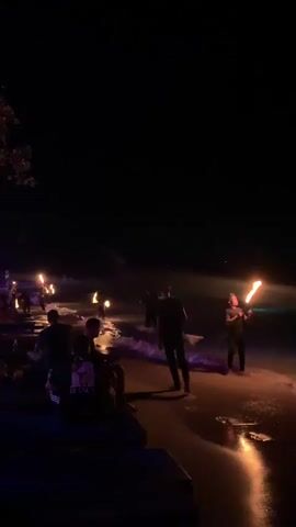 The video is a dimly lit nighttime scene on a beach. Several people are gathered, and some are performing with fire. In the foreground, silhouettes of people are seated on what appear to be benches or steps, facing away from the camera. To the right, several individuals are standing on the wet sand near the water's edge. Two of these individuals are holding torches with flames, one on the right of the frame and another further to the left. Another person is walking through the scene from right to left. In the background, more people are visible, and there are several more illuminated torches or fire-based lights spaced out along the shoreline. The overall atmosphere is dark with areas of intense orange light from the fires reflecting on the wet sand.