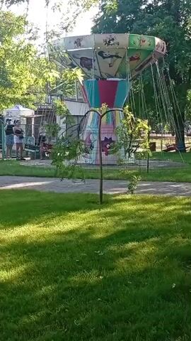 A carousel swing ride is in motion in a park. The ride has a central pole with colorful panels decorated with cartoon animals, and seats that hang from chains. Several people are visible on the ride, including children. The ride is surrounded by a fence, and beyond that, there is a grassy area with trees. The sun is shining, casting shadows on the grass. The video captures a few seconds of the ride's movement.