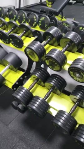 The video shows a variety of dumbbells of varying weights neatly arranged on a yellow rack in a well-equipped gym. The dumbbells are primarily black and have numerical labels indicating their weight. The rack holding the dumbbells is two-tiered, allowing for a wide range of weight options.

The camera slowly pans across the rack, highlighting the diversity of dumbbells. In the background, other pieces of gym equipment are visible, including a weight bench with a yellow and black frame and various weight machines.

The gym appears to be clean and well-maintained, with a black rubber floor and white walls. On the wall, a chart depicting human anatomy is visible.

The video has a static, observational quality, focusing on the equipment and gym environment rather than any action or interaction. The setting is clearly a professional or serious home gym setting.